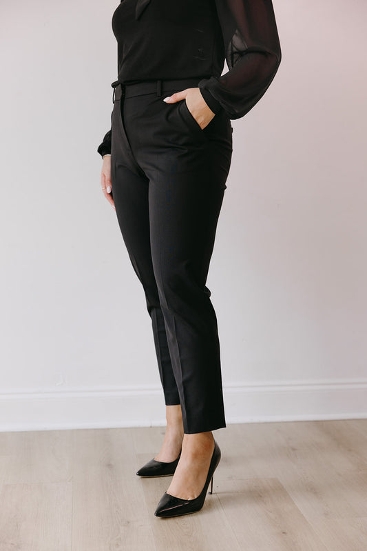 Person wearing a black outfit with pants and heels against a white wall.